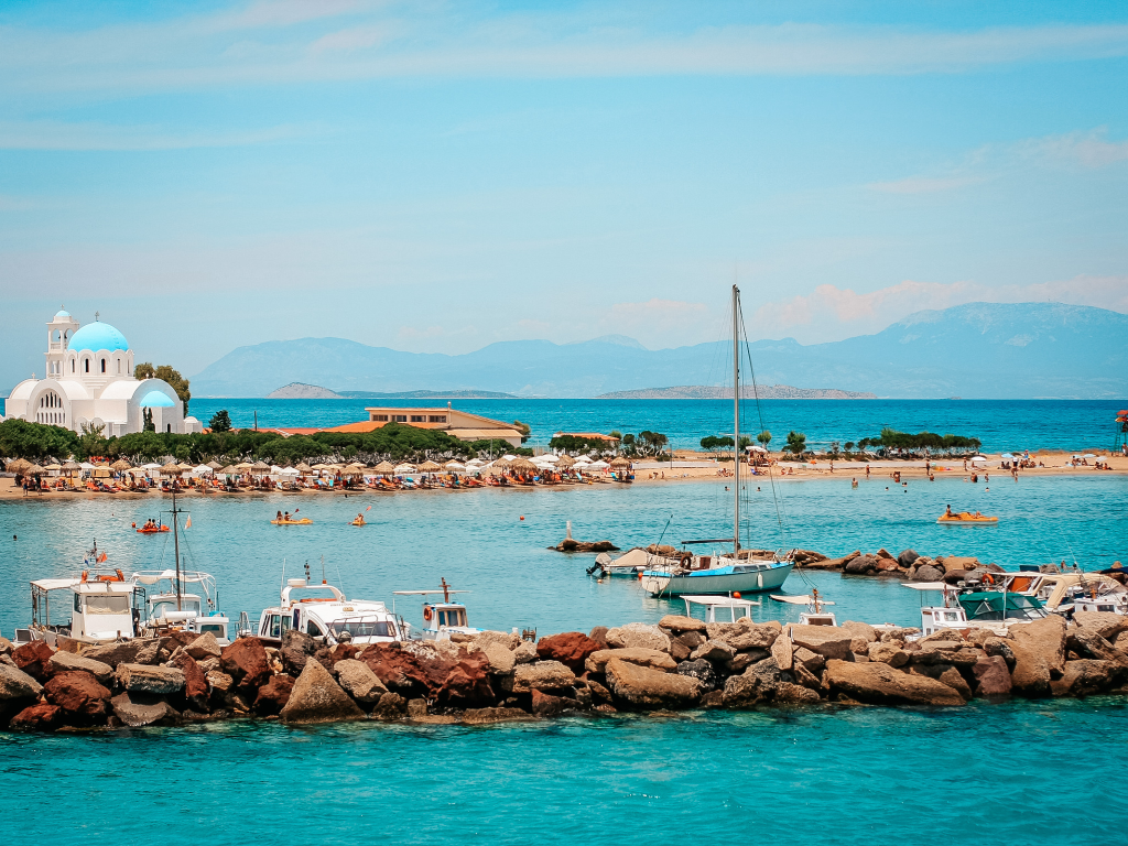 Barche ormeggiate nel porto di Skala, Agistri, con la Chiesa di Agioi Anargyroi che domina lo sfondo azzurro del Golfo del Saronico