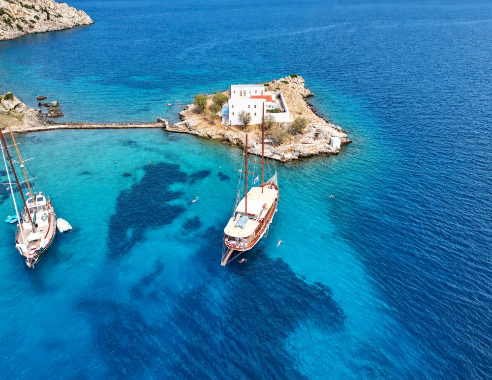 Vista dall'alto sulla chiesetta di Agios Emilianos su un piccolo isolotto di Symi, con due imbarcazioni ormeggiate nelle acque azzurre della baia.
