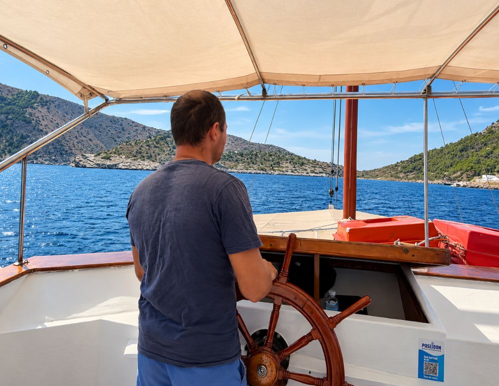Capitan Theodoros al timone dell'imbarcazione Poseidon durante l'escursione lungo la costa di Symi, con il mare e le colline sullo sfondo.
